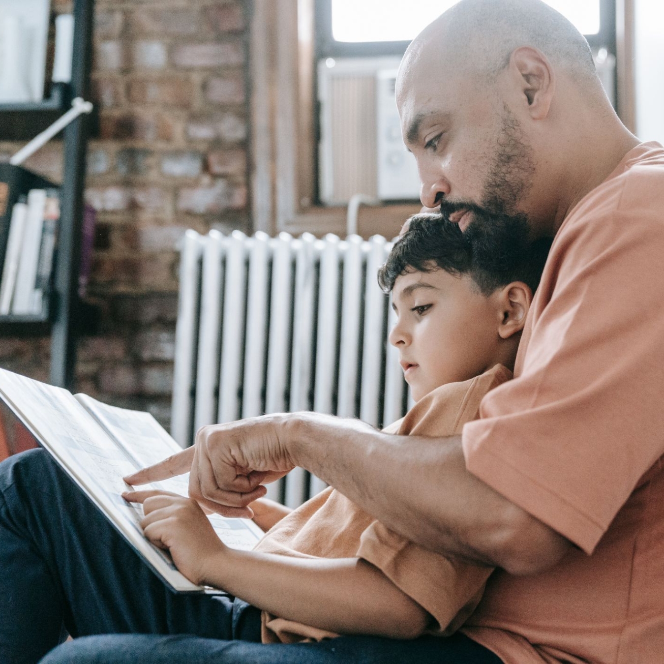 A father and son sitting on the floor reading a book together pointing at the words