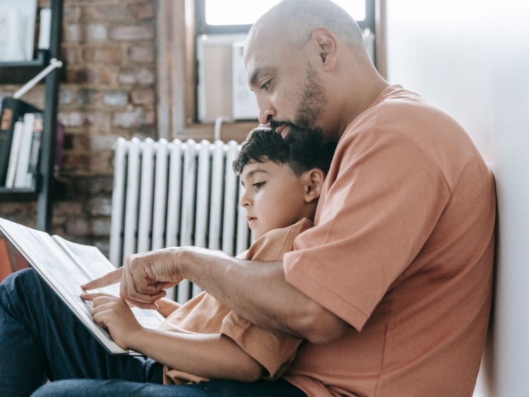 A father and son sitting on the floor reading a book together pointing at the words