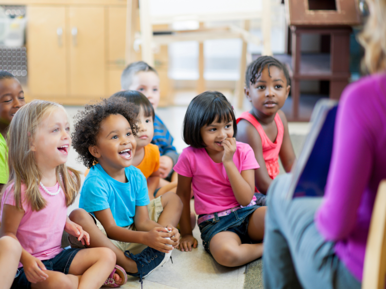 A group of small children sitting on the floor at pre school laughing at a book their teacher is reading