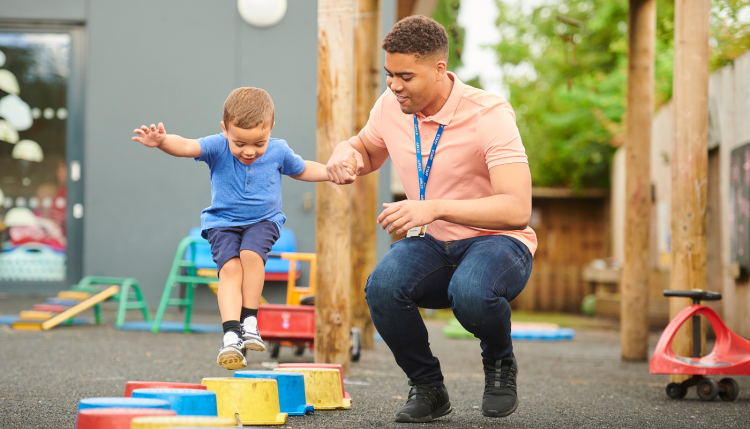 A young man with a lanyard on holding the hand of a young boy helping him jump over brightly coloured steps