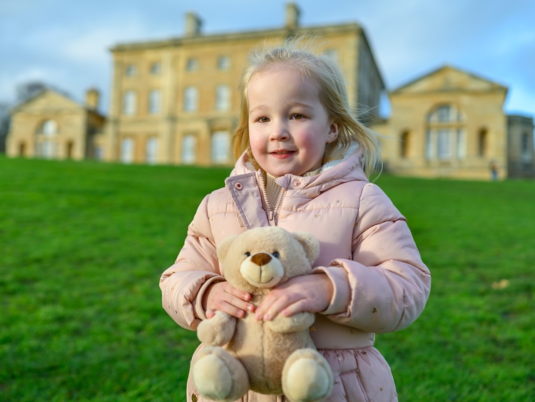 A little girl with blonde hair and pink coat holding a teddy in front of Cusworth Hall