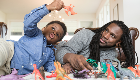 Dad with long dreadlocks and son lay on the floor playing with dinosaurs