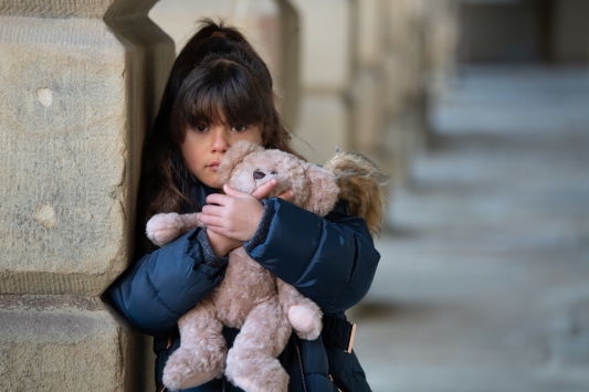 a little girl with dark brown hair clutching a teddy and looking sad, leaning against a stone pillar