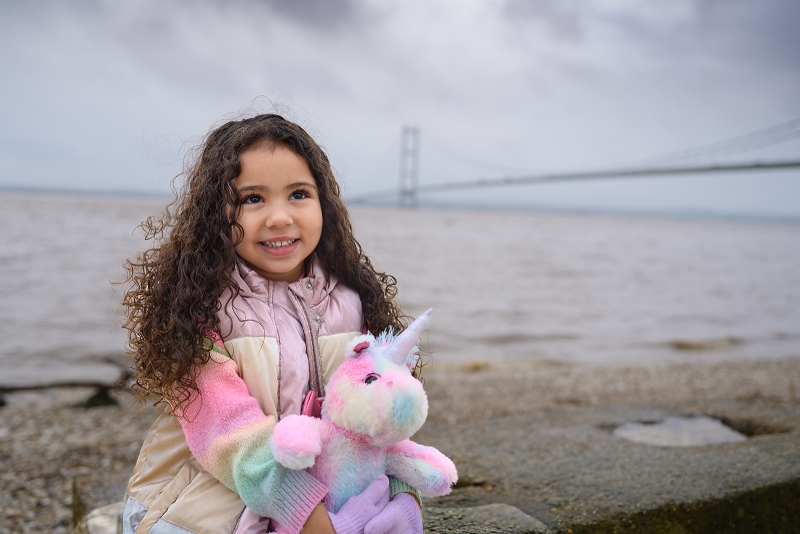 Little girl with long curly hair holding a rainbow unicorn with the humber bridge in background