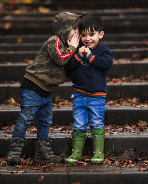 Two little boys whispering wearing wellies on steps in a park in autumn